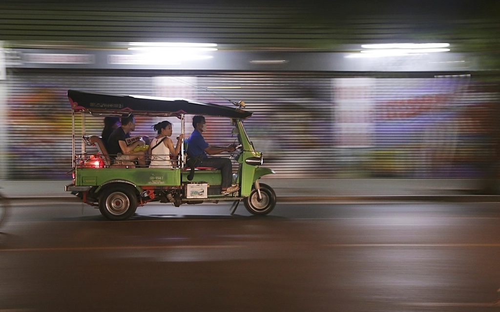 A green tuk-tuk carrying passengers down a Bangkok street at night